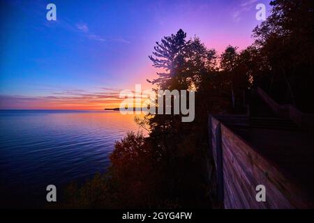 Sonnenaufgang aus Gold, rot, lila, rosa und blau über einem See oder Meer mit einem hölzernen Wanderweg auf der rechten Seite, der durch einen Wald schneidet Stockfoto