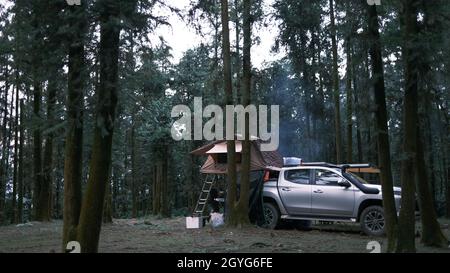 Camper genießen den alternativen Urlaub - Auto mit Zelt auf dem Dach und allein Mann Kochen im Wald. Abenteuer Reise Lifestyle Fernweh Stockfoto