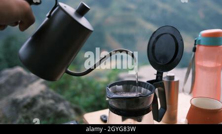 Der Barista bereitet beim Camping in der Natur Tropf zu, filtrierten Kaffee, gießt Kaffee mit heißem Wasser und Filterpapier über. Der Kaffee am Morgen ist großartig Stockfoto