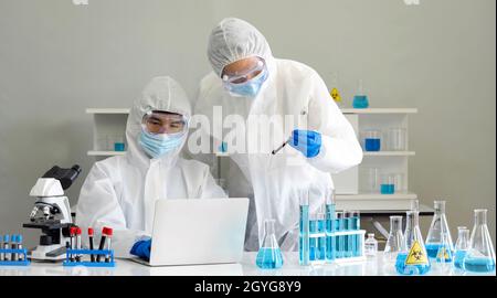 Epidemiologische Forscher in Virusschutzkleidung verwenden einen Laptop, um chemische Verbindungen im Internet zu erforschen. Coronavirus-Krankheit 2019 Stockfoto