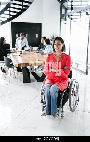 Porträt einer lateinischen Transgender-Frau im Rollstuhl und Menschen bei der Begegnung im Hintergrund im Büro in Lateinamerika Stockfoto