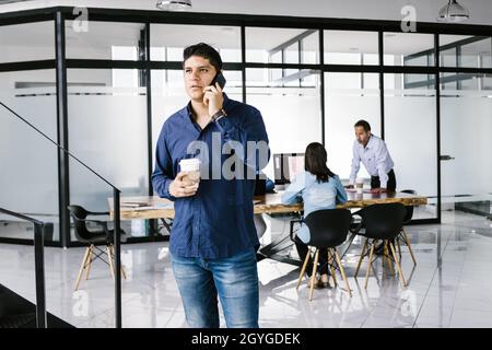 Porträt eines jungen hispanischen Geschäftsmannes mit einem Glas Kaffee, der vom Büro in Mexiko aus auf das Mobiltelefon ruft Stockfoto