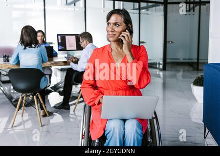 Die hispanische Transgender-Geschäftsfrau im Rollstuhl ist bei der Arbeit in Mexiko-Stadt mit einem Laptop an den Beinen beschäftigt und ruft am Handy an Stockfoto