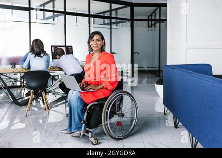 Lateinische Transgender-Frau, die in einem modernen Büro im Rollstuhl sitzt und einen Laptop mit Kollegen in Mexiko-Stadt benutzt Stockfoto