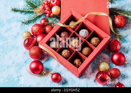 Pralinen in einer roten festlichen Schachtel dunkler Schokoladentrüffel auf weihnachtlichem Hintergrund mit Neujahrsspielbällen Tannenzweigen. Flacher imitierter Kopierraum. Stockfoto