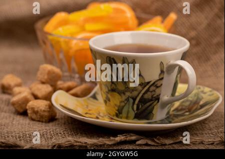 Bunte, geformte Marmeladenscheiben in einer Glasvase, Stücke braunen Rohrzuckers und eine Tasse Tee auf einer Untertasse, Nahaufnahme, selektiver Fokus Stockfoto