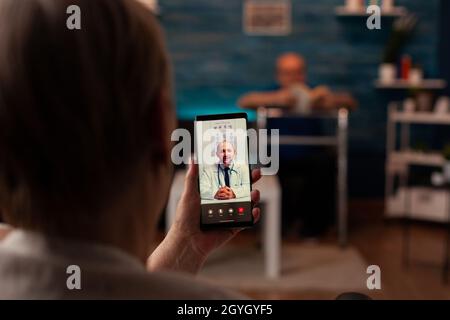 Alte Frau mit Behinderung nutzt Videoanruf für Telemedizin zu Hause. Gealterte Person, die mit dem Arzt zur Untersuchung einer Online-Fernkonferenz spricht, wobei das Smartphone im Rollstuhl sitzt Stockfoto