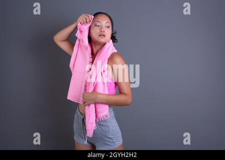 Junge Brünette chinesisches Mädchen in Sportswear und wischenden Schweiß mit Handtuch auf grauem Hintergrund. Stockfoto