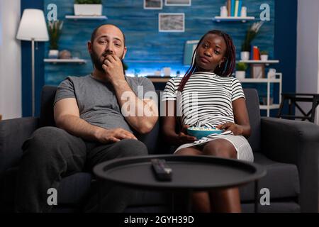 POV von interracial paar Fernsehen genießen Aktivität im Wohnzimmer. afroamerikanische Frau und kaukasischer Mann blicken auf die Kamera, während sie sich verkleben, entspannen und Popcorn essen Stockfoto
