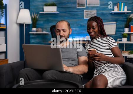 Moderne interracial paar mit Geräten und Technologie zu Hause. afroamerikanische Frau hält Smartphone, während kaukasischer Mann am digitalen Laptop arbeitet. Gemischtes Rennvolk mit Gadgets Stockfoto