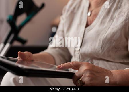 Nahaufnahme von nicht mehr erkennbaren, alten Händen, die den Tablet-Bildschirm zu Hause berühren. Ältere Frau, die ein digitales Gerät verwendet, während sie auf der Wohnzimmercouch mit Krücken zum Gehen sitzt. Ältere Erwachsene mit Technologie Stockfoto