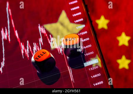 Würfel mit den Worten VERKAUFEN KAUFEN an den Rändern. Im Hintergrund die Grafik des Rückgangs des Aktienindex der chinesischen Unternehmen auf dem Smartphone-Bildschirm. Stockfoto