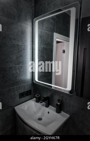 Moderne, einfarbige, graue Badezimmereinrichtung mit weißem Keramik-Eckwaschbecken oder Handwaschbecken unter einem großen gerahmten Spiegel Stockfoto