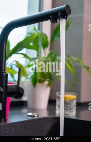 Nahaufnahme einer Küchenarmatur mit fließendem Wasser vor dem Hintergrund von grünen Topfpflanzen auf einer Fensterbank Stockfoto