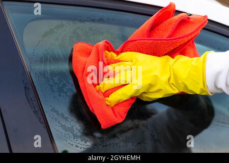 Eine Hand in einem gelben Gummihandschuh wischt das Glas eines Autos mit einem roten Mikrofaserlappen an einem warmen Herbsttag vom Schmutz ab. Nassreinigung. Selektiver Fokus. Clos Stockfoto