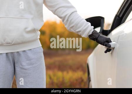 Ein Mann in einem weißen Pullover und schwarzen Handschuhen öffnet die Autotür, um ihn an einem warmen Herbsttag zu stehlen. Selektiver Fokus. Nahaufnahme Stockfoto