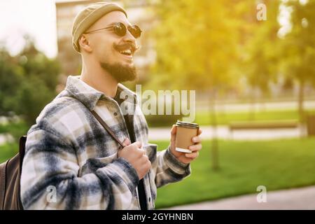 Ein gutaussehender, fröhlich lächelnder, bärtiger Mann trinkt vor der Straße Kaffee, während er im Stadtpark spaziert Stockfoto