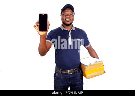 Junger Mann mit Mütze, der auf weißem Hintergrund steht und das Mobiltelefon zeigt und lächelnd einen Karton in der Hand hält. Stockfoto