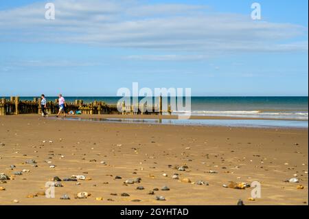 Ein Paar, das mit seinem Hund neben einer hölzernen Groyne in der Sonne am Strand von Mundesley, Norfolk, England, läuft. Stockfoto