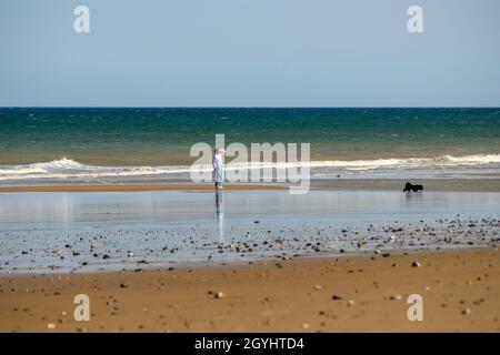 Eine Frau geht mit ihrem Hund an einem ansonsten leeren Strand von Mundesley bei strahlendem Sonnenschein, Norfolk, England. Stockfoto