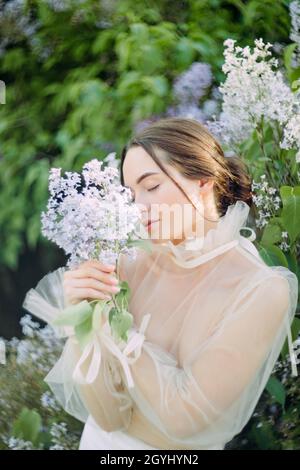 Porträt einer jungen Frau im Garten mit einem Fliederzweig in der Hand vor dem Hintergrund blühender Sträucher. Stockfoto