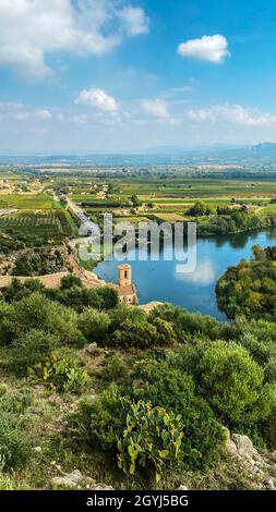 Der Ebro-Fluss, der durch Miravet, Spanien, führt und den Glockenturm seiner Kirche und die Bergkette Serra de Cardo und Els Ports im Hintergrund beleuchtet Stockfoto