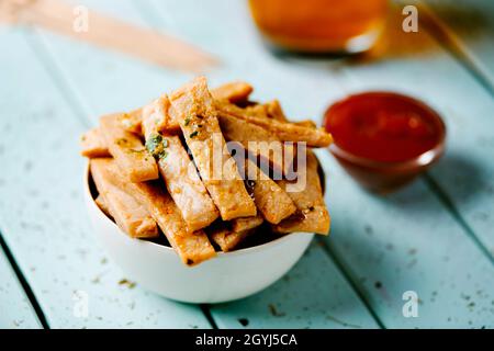 Nahaufnahme von einigen frittierten Hähnchenstreifen in einer Schüssel, die auf einem blauen Tisch neben einer kleinen Schüssel mit Ketchup platziert wird Stockfoto