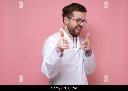 Ein lustiger Arzt in einem weißen Mantel scherzt und lächelt und zeigt auf jemanden mit den Fingern. Rosa Hintergrund. Stockfoto