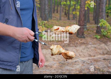 Gegrilltes Fleisch in der Hand eines Mannes in der Natur im Lager. Shish Kebab Braten auf dem Grill im Freien. Hühnerfleisch wird gebraten. Stockfoto