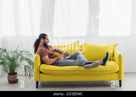 Seitenansicht des hispanischen Mannes, der zu Hause auf der gelben Couch sitzt und einen Laptop benutzt Stockfoto