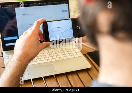 Broker mit Smartphone, um Fotos von Charts am Computer zu machen, während er am Tisch sitzt und in den Aktienmarkt im Home Office voller Pflanzen investiert. Stockfoto