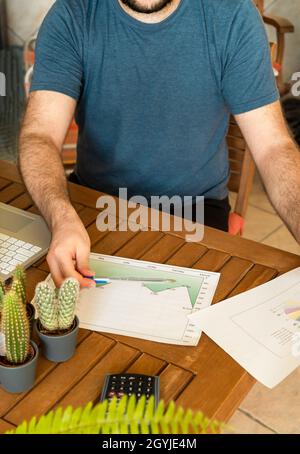 Bärtiger junger Broker, der Charts mit einem Computer analysiert, während er vom Outdoor-Heimbüro voller Pflanzen arbeitet. Stockfoto