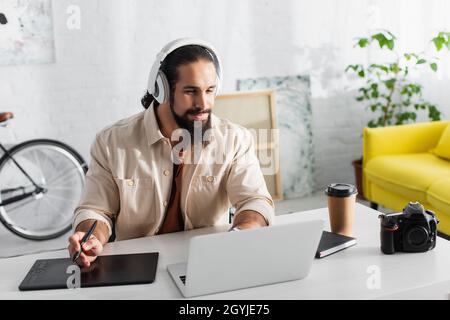 hispanischer Designer mit Kopfhörern, der in der Nähe von Laptop, Grafiktablett und Digitalkamera arbeitet Stockfoto