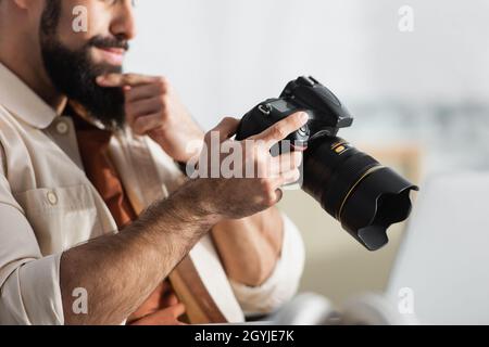 Beschnittene Ansicht eines durchdachten Mannes, der den Bart berührt, während er die Digitalkamera zu Hause hält Stockfoto