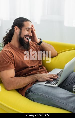 Aufgeregter hispanischer Mann lacht, während er zu Hause einen Laptop auf der Couch benutzt Stockfoto