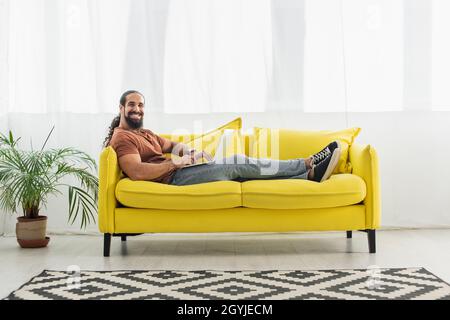 Fröhlicher hispanischer Mann, der die Kamera anlächelt, während er einen Laptop auf dem gelben Sofa benutzt Stockfoto