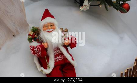 Bringen Sie den Weihnachtsmann mit einer Geschenktüte auf seiner Schulter und einem Holzschlitten zum Spielzeug. Eine Puppe Weihnachtsmann mit Brille und weißem Bart sitzt im Schnee. Stockfoto