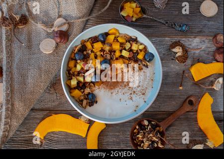 Gesunde Frühstückschale für Herbst- und Wintersaison mit hausgemachtem Müsli, Äpfeln, Kürbis, Blaubeeren und Joghurt auf Holztisch. Ansicht von oben Stockfoto