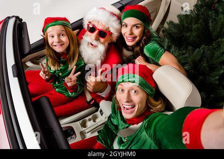 Foto von ziemlich niedlichen santa claus Assistenten fahren Auto zeigt V-Zeichen Daumen nach oben isoliert weiße Farbe Hintergrund Stockfoto