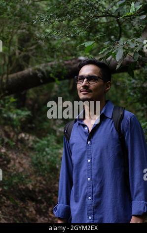 Junger inder mit Brille, der im Wald wegschaut. Überlebender, der denkt, während er in der Nähe eines umgestürzten Baumes steht Stockfoto