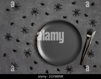 Halloween Tischunterlage mit schwarzem Geschirr und Spinnen auf dunkelgrauem Hintergrund. Leere schwarze Platte für die Konstruktion. Platz zum Kopieren, Draufsicht, flach liegend. Stockfoto