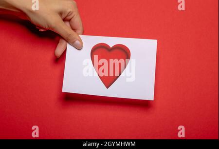 Weibliche Hand hält ein Stück Papier mit ausgeschnittener Herzform als Liebessymbol über starkem roten Hintergrund Karton Stockfoto