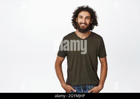 Gutaussehender, lächelnder Typ aus dem Nahen Osten mit langem Bart und lockigem Haar, der in legerer Kleidung vor weißem Hintergrund steht Stockfoto
