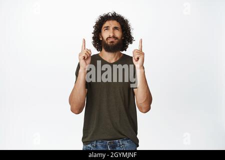 Zweifelhafter Mann, der die Finger nach oben zeigt, mit zögerlichem, skeptischem Gesichtsausdruck auf weißem Hintergrund steht Stockfoto