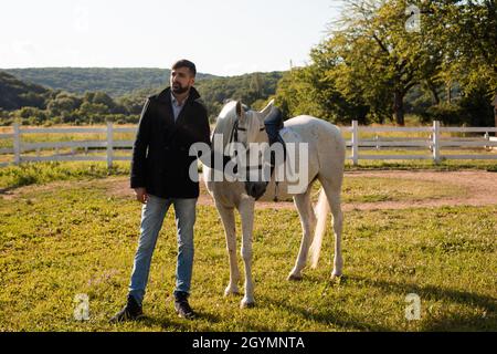 Der junge Mann in einer schwarzen Jacke steht mit einem weißen Pferd auf der Ranch. Stockfoto