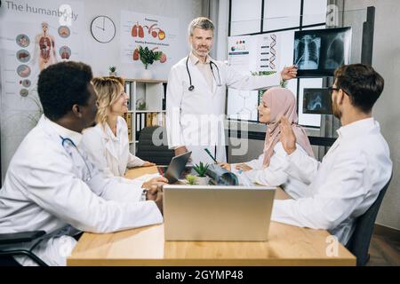 Gruppe von multirassischen Ärzten mit einem im Rollstuhl, die sich im Sitzungssaal versammeln, um die Ergebnisse der Röntgenaufnahme zu untersuchen. Kaukasischer Senior Therapeut lächelt und blickt auf die Kamera, während er auf den Scan zeigt. Stockfoto