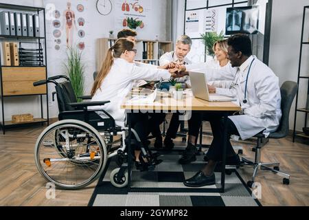 Multirassische Ärzte, bei denen eine Frau im Rollstuhl ihre Hände im Kreis zusammenhält, während sie sich erfolgreich im Büro trifft. Konzept der Medizin, Wissenschaft und Zusammenarbeit. Stockfoto
