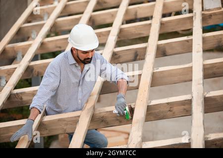 Der junge Mann in einem Schutzhelm und Fäustlingen arbeitet mit einem Pinsel auf dem Dach. Er beendet den Holzrahmen auf dem Dach Stockfoto