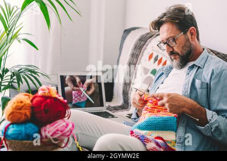 Ein erwachsener Mann zu Hause folgt Online-Strickanleitung, um sich zu entspannen und die Freizeit zu genießen, die auf dem Sofa sitzt - männliche Menschen stricken mit com Stockfoto