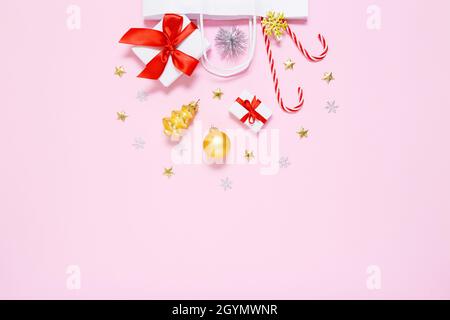 Weihnachten, Neujahrskomposition. Weiße Papiertüte mit Geschenken, süßen Bonbons, Weihnachtsdekorationen, Goldkugeln, Glitzer-Konfetti-Sternen auf Pastell-P Stockfoto